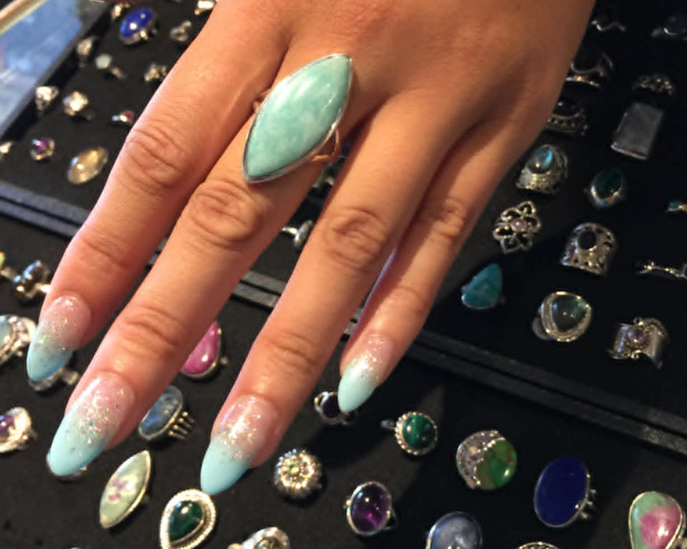 Manicured hand wearing a turquoise blue crystal ring with a display of various rings in the background.