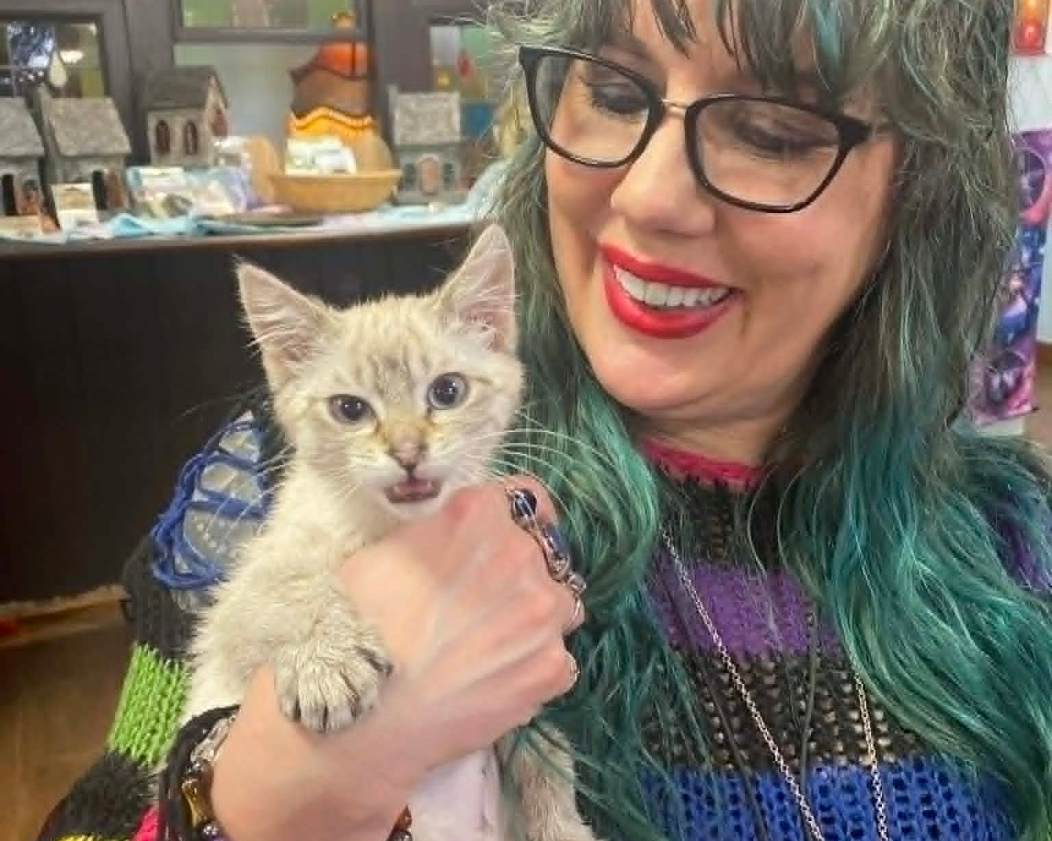 Shop employee with teal hair and a colorful sweater holding a small kitten at our store.