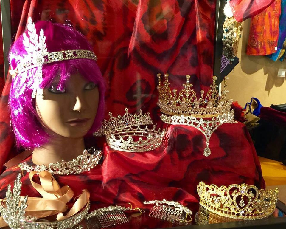 Mannequin head with purple wig wearing a jeweled headpiece, surrounded by crystal tiaras in a display case.