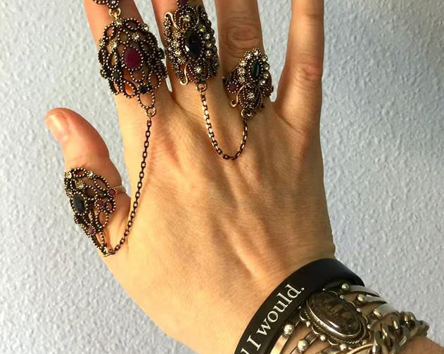 Hand wearing multiple ornate rings and bracelets against a plain background.