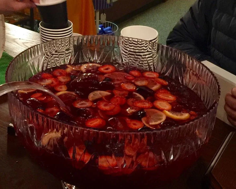 Punch bowl filled with strawberry lemonade and sliced fruits with a ladle on a table surrounded by people at a shop party.