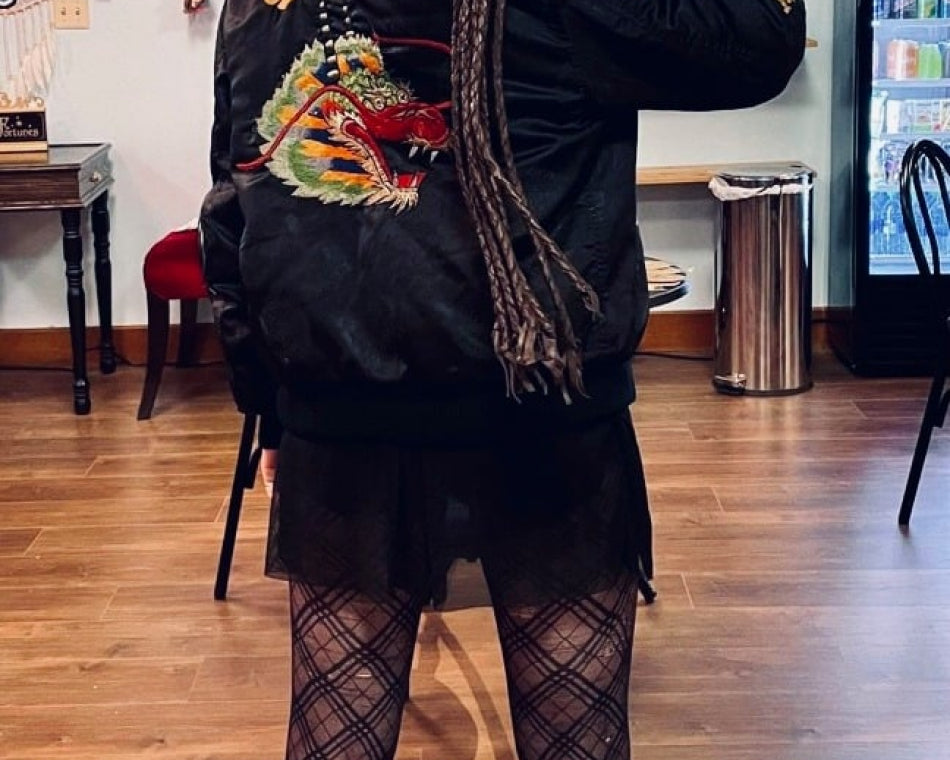 Shop regular wearing a black jacket with an embroidered dragon, fishnet tights, and boots, holding a leather whip.