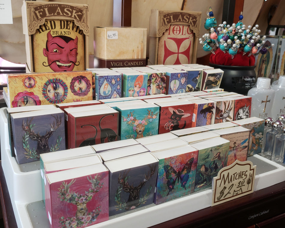 The image features charming and mystical designs on small match boxes, displayed on a shelf with a flask in the background.