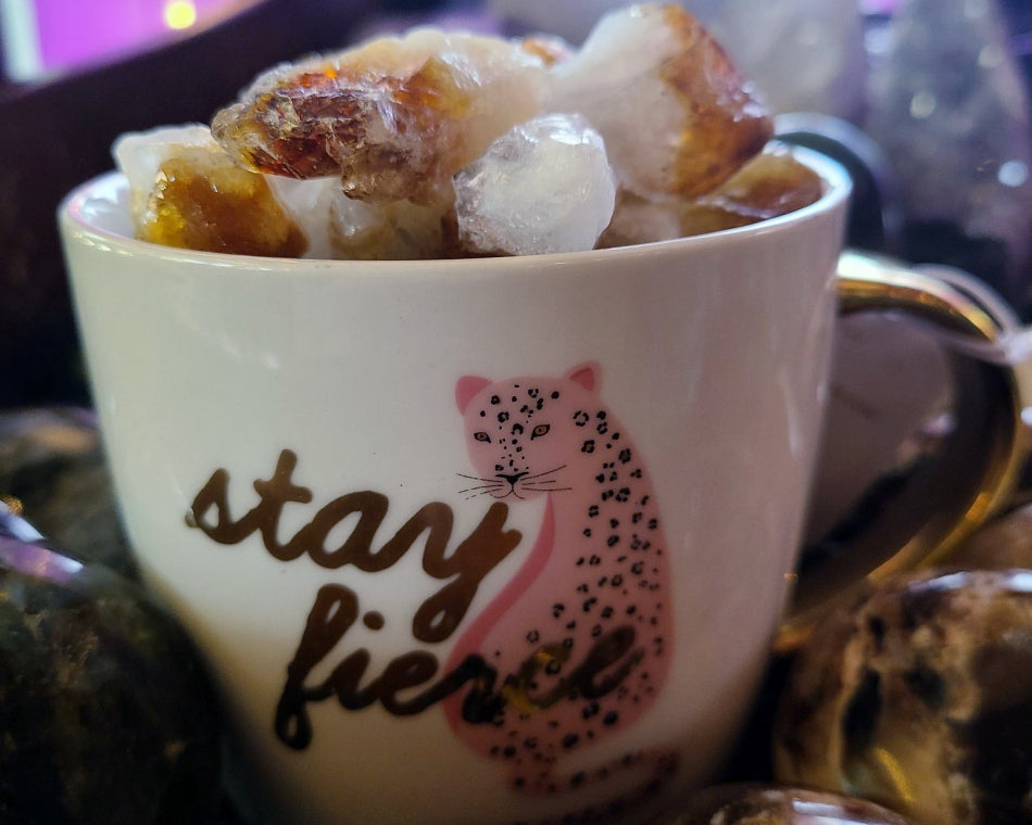 A white and gold mug with a pink jaguar and the words "Stay Fierce." Citrine crystals fill the mug.