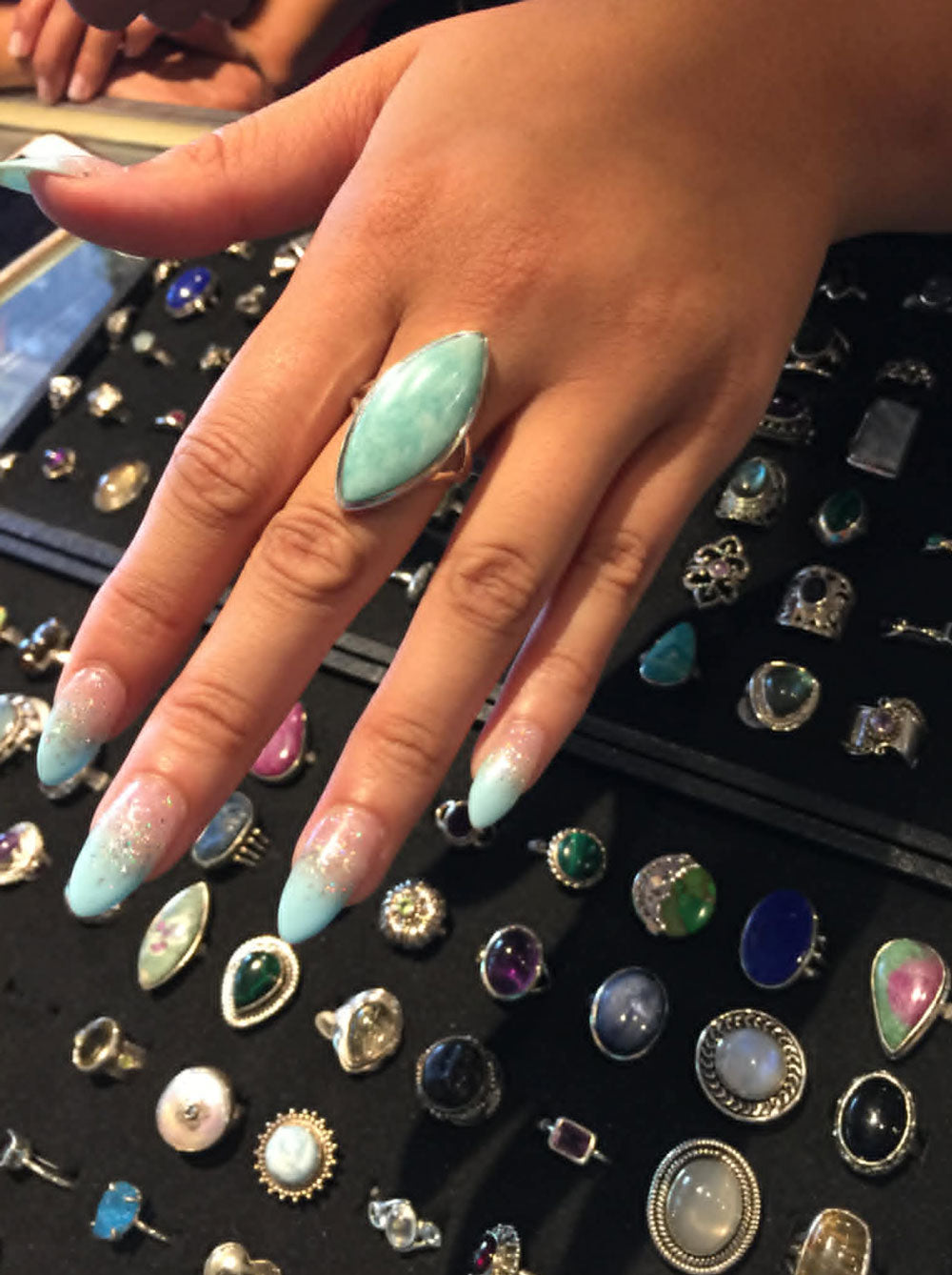 Manicured hand wearing a turquoise blue crystal ring with a display of various rings in the background.