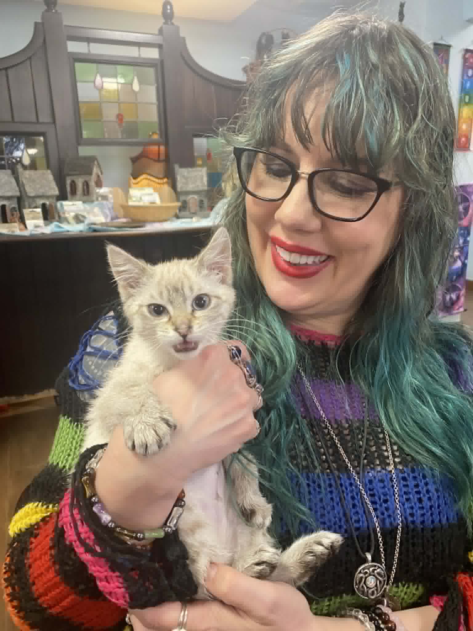 Shop employee with teal hair and a colorful sweater holding a small kitten at our store.