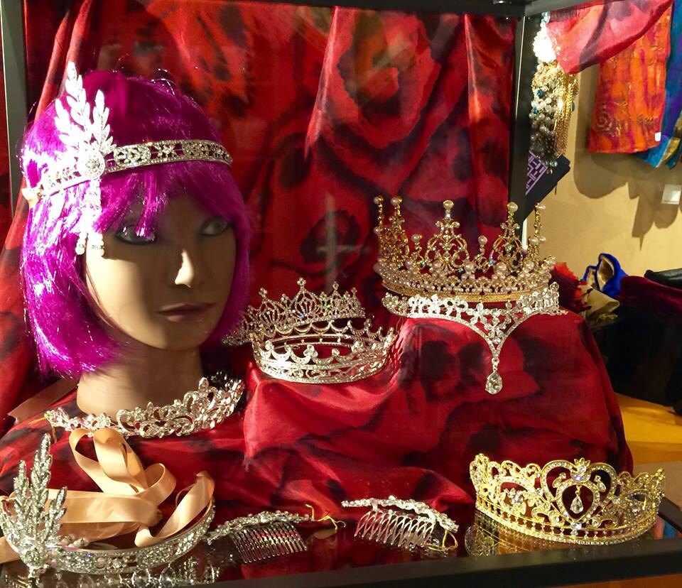 Mannequin head with purple wig wearing a jeweled headpiece, surrounded by crystal tiaras in a display case.
