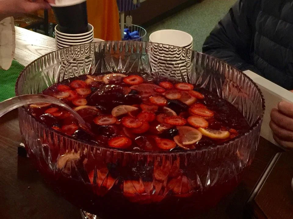 Punch bowl filled with strawberry lemonade and sliced fruits with a ladle on a table surrounded by people at a shop party.