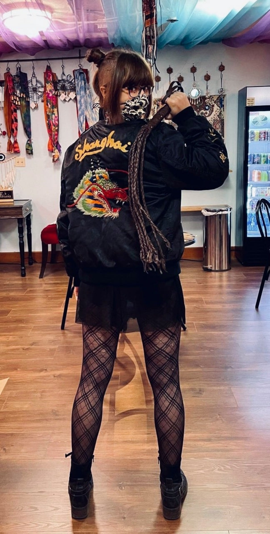 Shop regular wearing a black jacket with an embroidered dragon, fishnet tights, and boots, holding a leather whip.