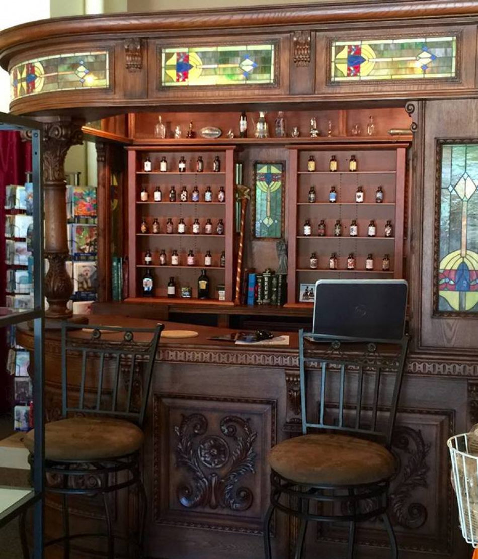 Wooden bar with stained glass panels and shelves filled with perfume bottles, surrounded by chairs.